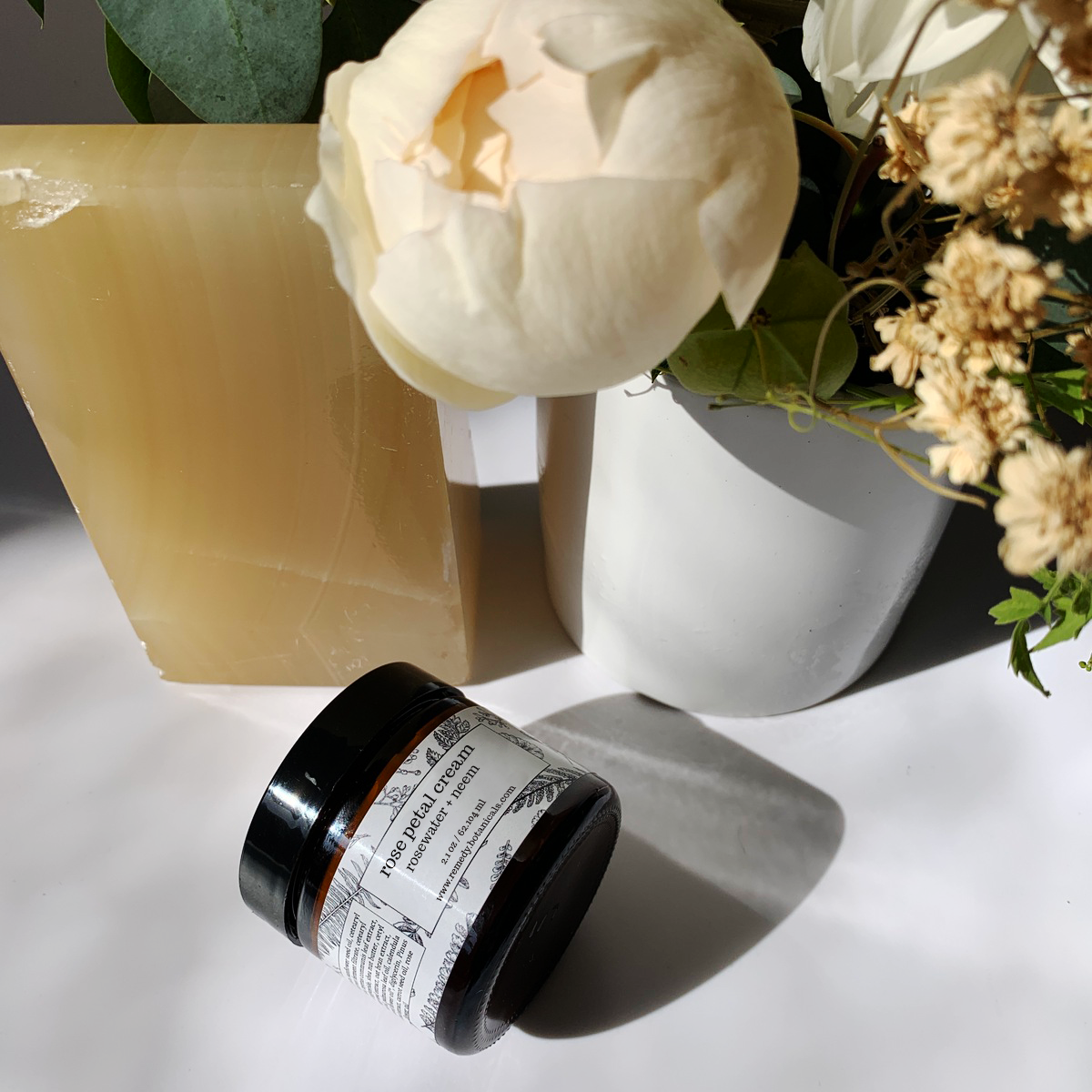 Top view of a bottle of Rose Water Cream beside bright orange bars, with an assortment of vibrant florals in the background, creating a fresh and elegant composition.