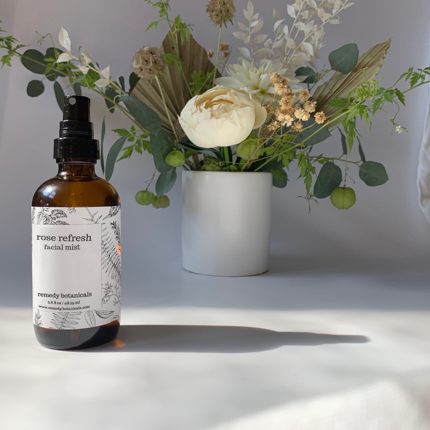 A bottle of Rose Refresh Facial Mist stands on the left side, centered next to a variety of flowers arranged in a white vase, set against a soft gray background. The arrangement evokes freshness and natural beauty.