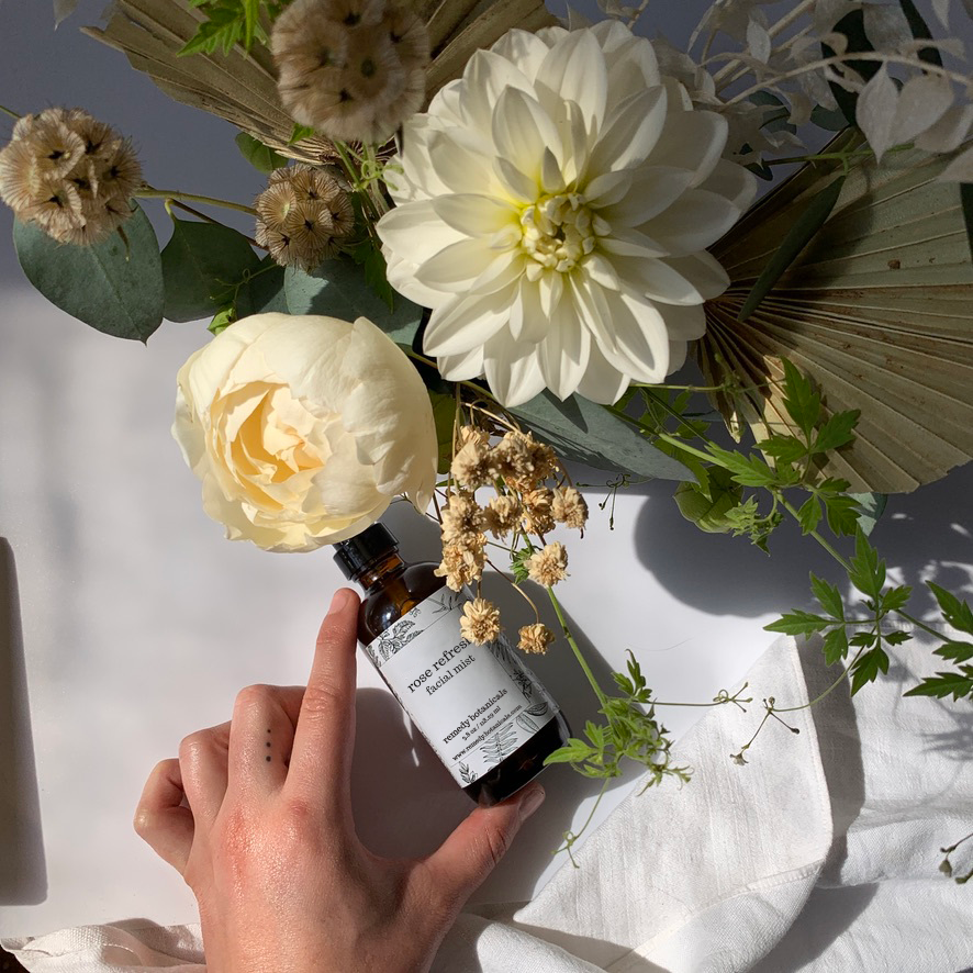 Top view of a bottle of Rose Refresh Facial Mist laying down, surrounded by various white flowers and green leaves, held delicately in hand. The scene exudes freshness and natural beauty.